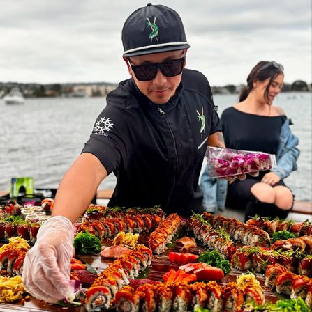 Sushi chef arranging a vibrant sushi spread on a wooden board at a waterfront event — rows of colorful rolls, sashimi, seaweed salad and orchid garnishes.
