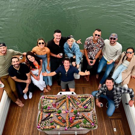 Aerial view of a smiling group of friends on a boat deck over green water, gathered around an elaborate sushi platter for a casual party.
