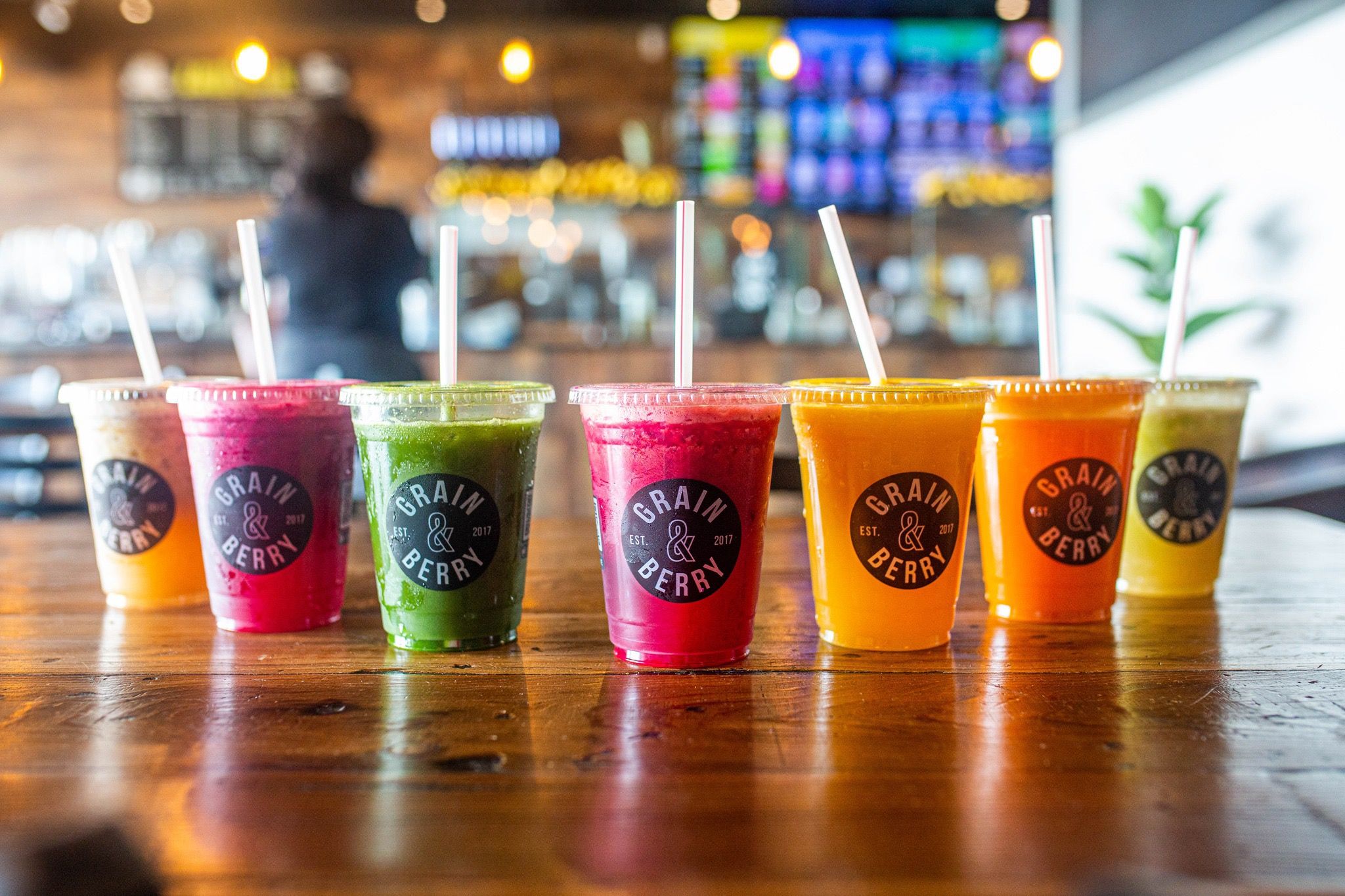 Row of colorful fruit and vegetable smoothies in clear plastic cups with lids and straws on a wooden table inside a bright modern juice bar, featuring pink, green, orange and yellow blends.