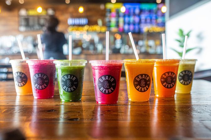 Row of colorful fruit and vegetable smoothies in clear plastic cups with lids and straws on a wooden table inside a bright modern juice bar, featuring pink, green, orange and yellow blends.