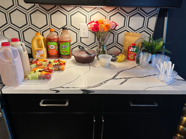 Bright kitchen countertop brunch spread on a marble island with hexagon tile backsplash: fresh fruit cups, juice and smoothie jugs, cocktail mix, clear cups and utensils, and a vase of roses.