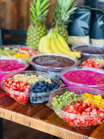 Vibrant acai and pitaya smoothie bowls on a wooden table topped with fresh strawberries, blueberries, kiwi, mango and pineapple, with bananas and whole pineapples in the background.