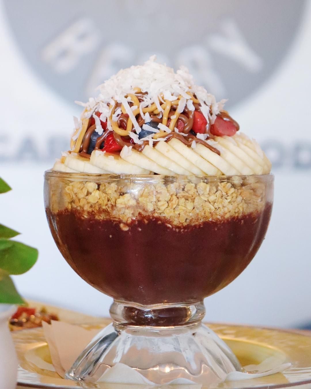 Acai bowl in a glass parfait dish with deep purple acai base, crunchy granola, fanned banana slices, peanut-butter and chocolate drizzle, fresh berries and shredded coconut