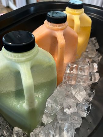 Three colorful plastic juice jugs — green, orange, and yellow — chilled on ice in a black serving tub
