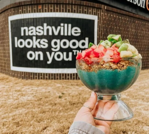 Hand holding a vibrant blue smoothie bowl topped with granola, diced strawberries and kiwi, posed in front of a brick wall mural that reads Nashville looks good on you.