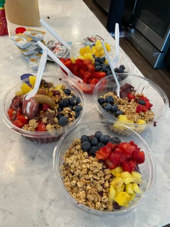 Colorful acai bowls in clear to-go containers topped with granola, blueberries, diced pineapple and strawberries, with white plastic spoons on a white marble kitchen countertop.