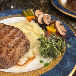 Gourmet grilled steak with creamy mashed potatoes, sautéed mushrooms, roasted peppers and microgreens on a blue and gold dinner plate.