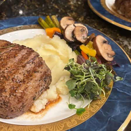 Gourmet grilled steak with creamy mashed potatoes, sautéed mushrooms, roasted peppers and microgreens on a blue and gold dinner plate.