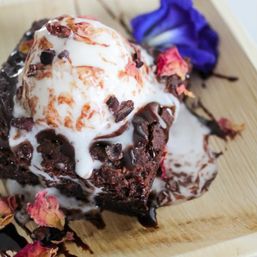 Close-up of an indulgent warm chocolate brownie topped with melting vanilla ice cream, chocolate drizzle and cacao nibs, garnished with dried rose petals and a purple edible flower on a wooden serving board.