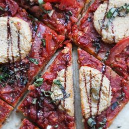 Close-up of sliced gourmet vegan pizza topped with chunky tomato sauce, dollops of cashew ricotta, chopped basil and a balsamic glaze drizzle