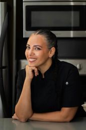 Smiling female chef with braided hair and nose ring in a black chef coat leaning on a counter in a modern kitchen with stainless steel oven and refrigerator.