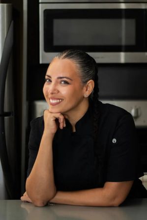 Smiling female chef with braided hair and nose ring in a black chef coat leaning on a counter in a modern kitchen with stainless steel oven and refrigerator.
