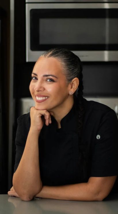 Smiling female chef with braided hair and nose ring in a black chef coat leaning on a counter in a modern kitchen with stainless steel oven and refrigerator.