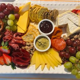 Vibrant charcuterie board with wedges of cheddar and brie, sliced cheese, cured meats and pepperoni, green and red grapes, strawberries, assorted crackers and pretzels, dried apricots, candied nuts and two dipping sauces.