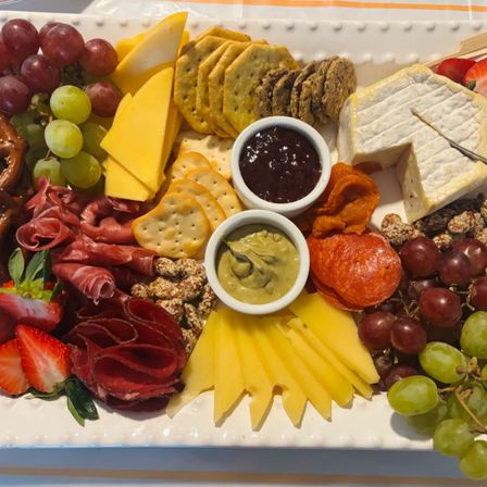 Vibrant charcuterie board with wedges of cheddar and brie, sliced cheese, cured meats and pepperoni, green and red grapes, strawberries, assorted crackers and pretzels, dried apricots, candied nuts and two dipping sauces.