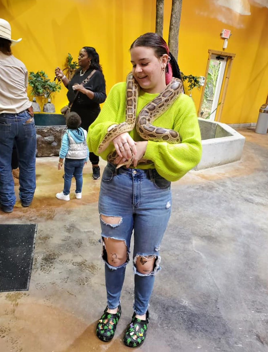 Smiling visitor in a neon green sweater holding a large python draped around their shoulders at an indoor reptile exhibit with yellow walls and other visitors in the background.