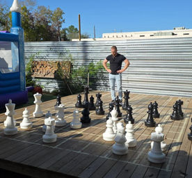 Giant outdoor chess set on a wooden deck with oversized black and white pieces on a painted grid, a man in a black shirt and light jeans studying the board, corrugated metal fence and inflatable bounce house under a clear blue sky.
