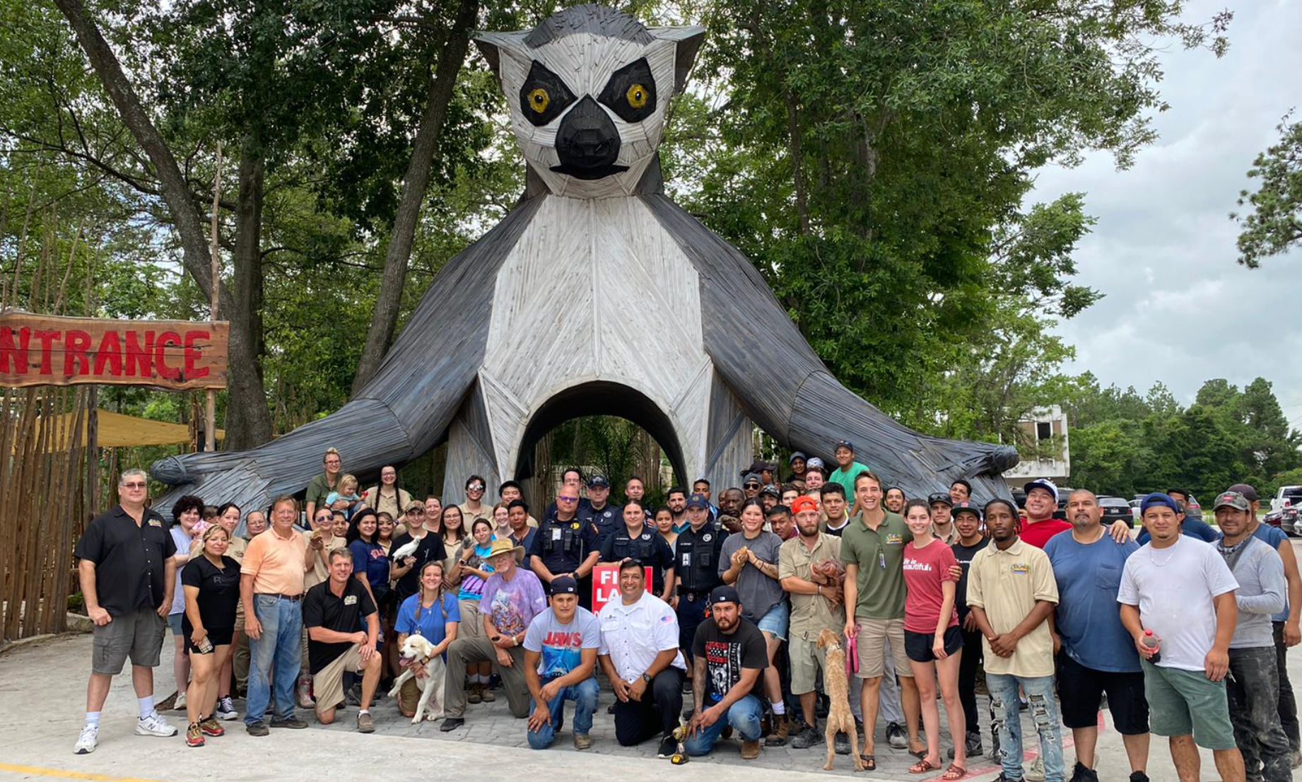 Large group of people posing beneath a towering wooden lemur sculpture that forms an archway entrance at an outdoor wooded attraction, trees and a red "ENTRANCE" sign visible.