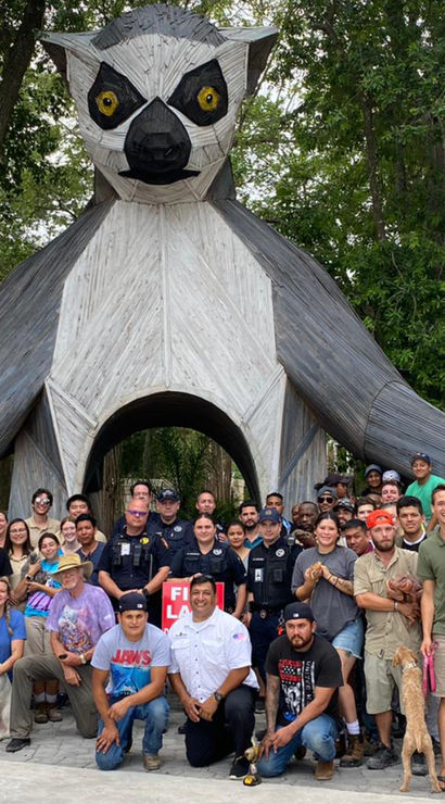 Large group of people posing beneath a towering wooden lemur sculpture that forms an archway entrance at an outdoor wooded attraction, trees and a red "ENTRANCE" sign visible.