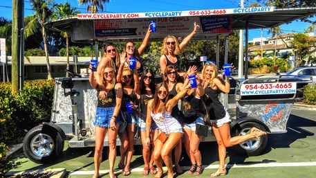 Group of women in summer outfits posing and celebrating on a pedal-powered party bike, holding blue cups and smiling under palm trees on a sunny day