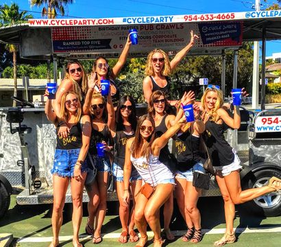 Group of women in summer outfits posing and celebrating on a pedal-powered party bike, holding blue cups and smiling under palm trees on a sunny day