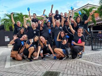 Cheerful team photo of a large group in matching black shirts waving and holding blue cups in a palm‑lined outdoor plaza with tiled roofs — lively celebration or company outing.