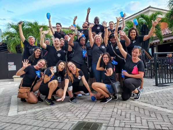 Cheerful team photo of a large group in matching black shirts waving and holding blue cups in a palm‑lined outdoor plaza with tiled roofs — lively celebration or company outing.