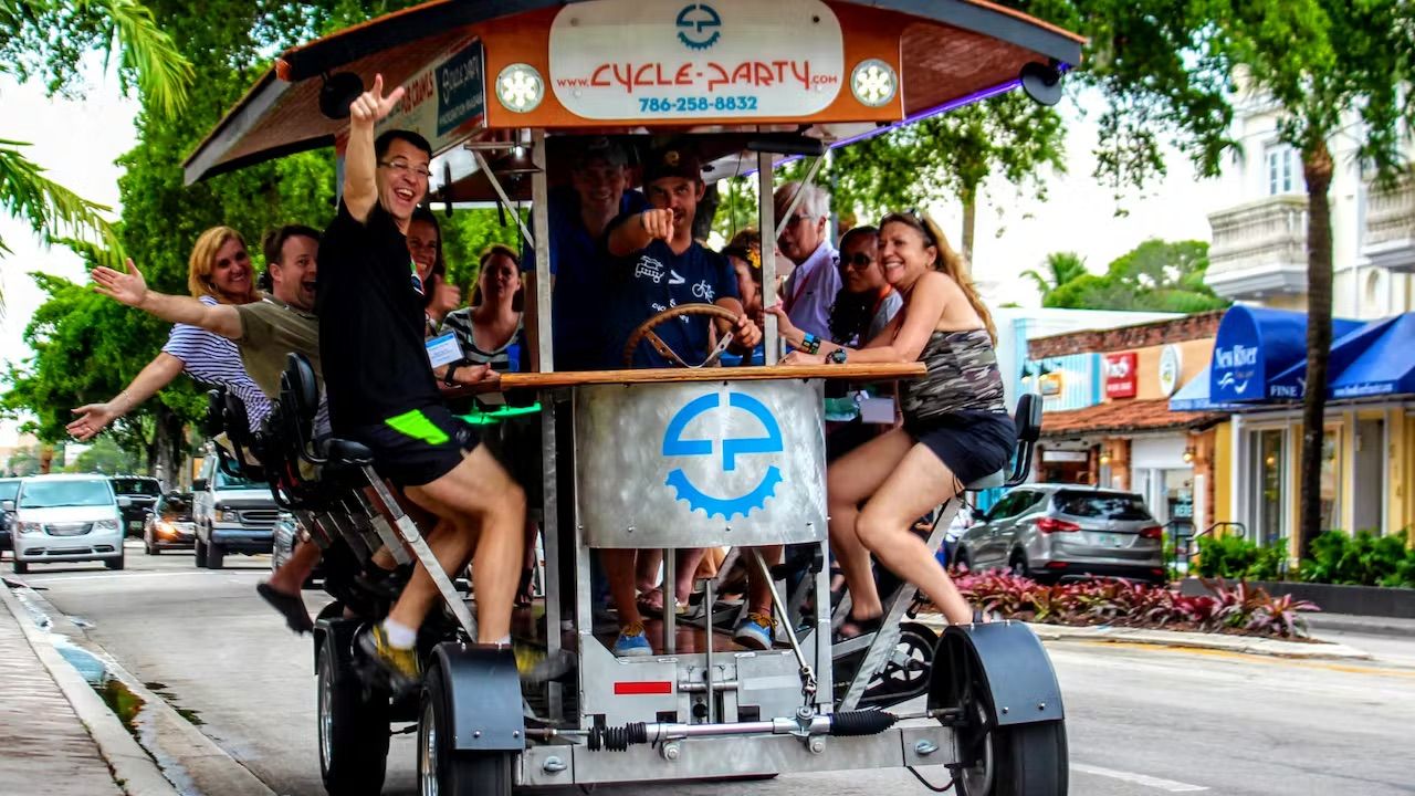 Smiling adults cheering and waving on a pedal-powered party bike with a wooden canopy, pedaling past palm trees and storefronts on a lively downtown street.