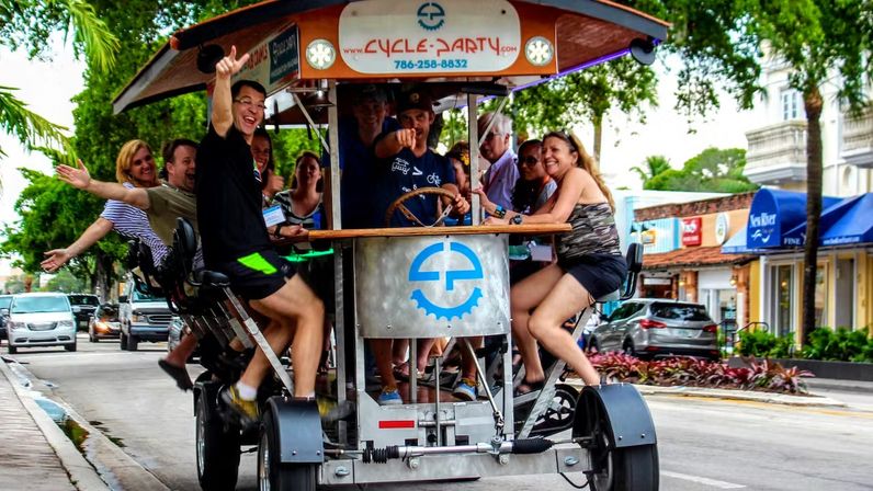 Smiling adults cheering and waving on a pedal-powered party bike with a wooden canopy, pedaling past palm trees and storefronts on a lively downtown street.