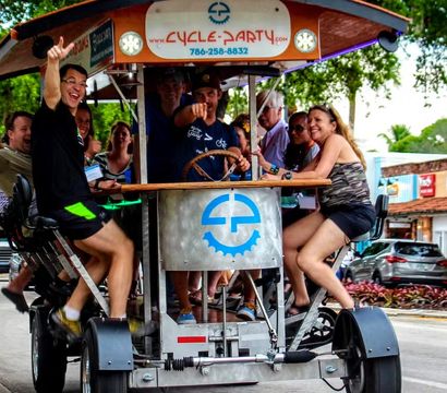 Smiling adults cheering and waving on a pedal-powered party bike with a wooden canopy, pedaling past palm trees and storefronts on a lively downtown street.