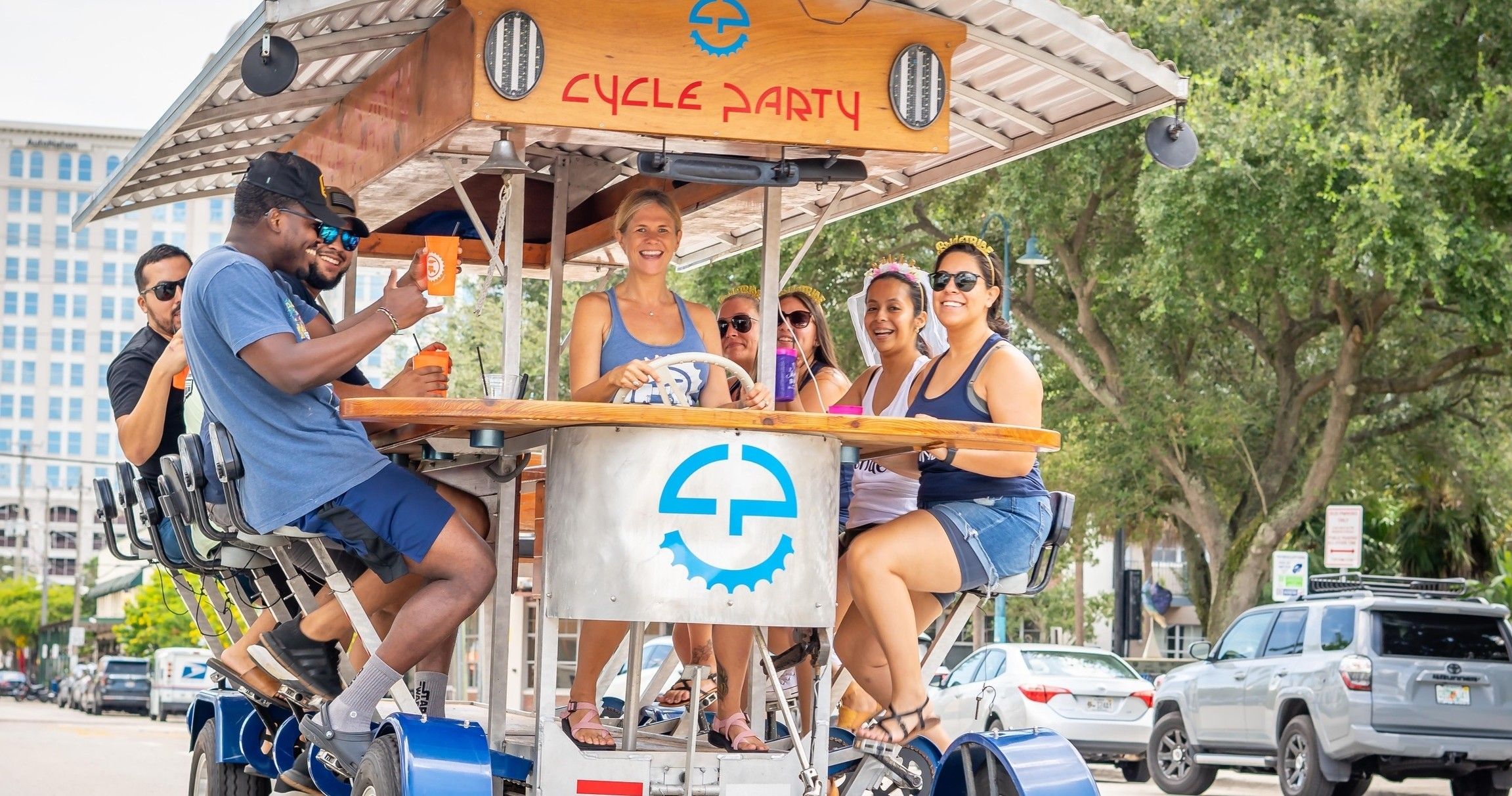 Smiling group of adults on a pedal-powered party bike riding down a downtown street, seated around a central bar table with drinks.