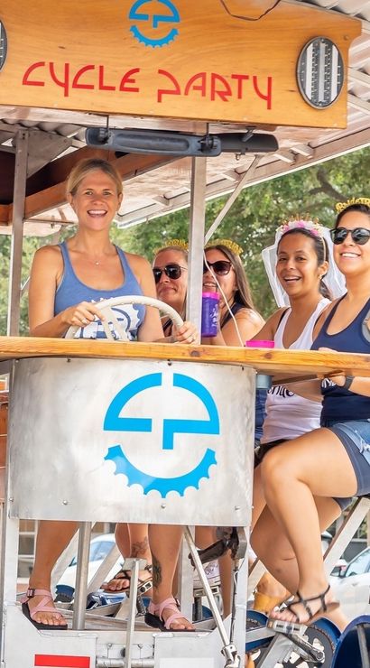 Smiling group of adults on a pedal-powered party bike riding down a downtown street, seated around a central bar table with drinks.