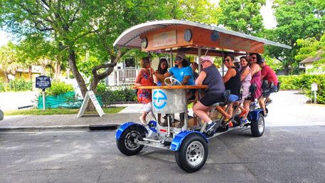 Group of people riding a colorful pedal-powered party bike with a wooden canopy and bar-style seats down a sunny tree-lined street, enjoying an outdoor group cycling tour.