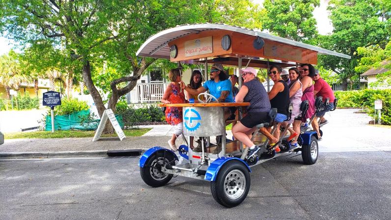 Group of people riding a colorful pedal-powered party bike with a wooden canopy and bar-style seats down a sunny tree-lined street, enjoying an outdoor group cycling tour.