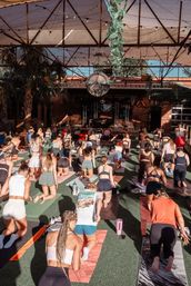 Sunlit outdoor group yoga class on an urban covered patio — dozens on colorful mats kneeling toward a stage under string lights, disco ball, and hanging plants.