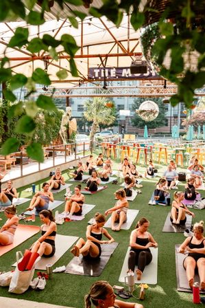 Vibrant urban rooftop fitness class on artificial turf under a canopy — dozens practicing core exercises on yoga mats amid hanging plants, colorful bar seating, palm trees and a disco ball.