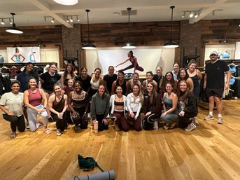 Large group of people in athletic wear smiling for a post-workout group photo inside an activewear retail store — wooden floors, hanging pendant lights, apparel racks and a yoga mural in the background.