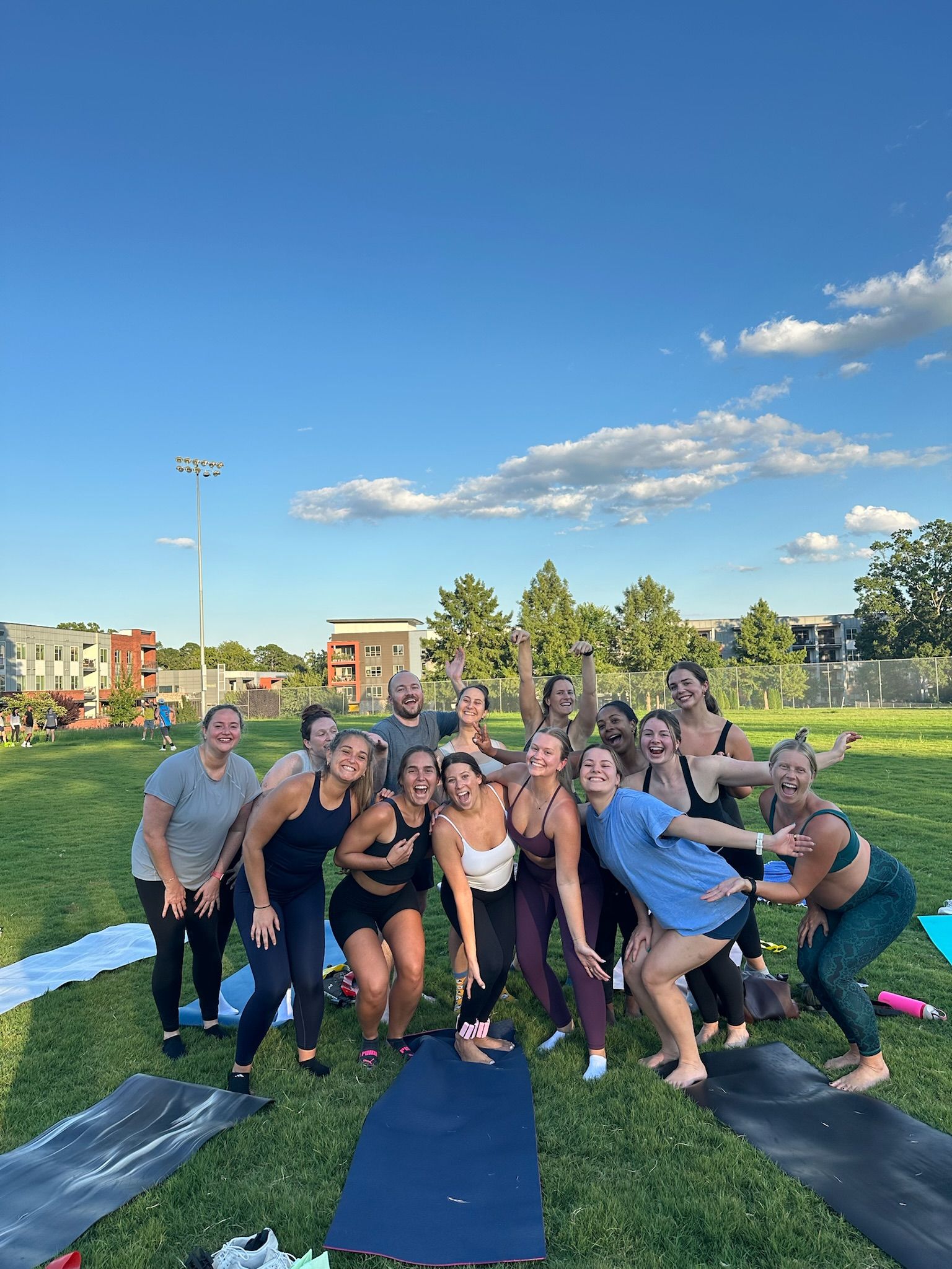 Energetic group of people in athletic wear posing and laughing after an outdoor yoga/fitness class on a sunny urban park lawn with yoga mats, apartment buildings and a blue sky with scattered clouds.