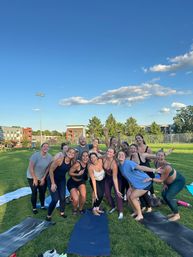 Energetic group of people in athletic wear posing and laughing after an outdoor yoga/fitness class on a sunny urban park lawn with yoga mats, apartment buildings and a blue sky with scattered clouds.