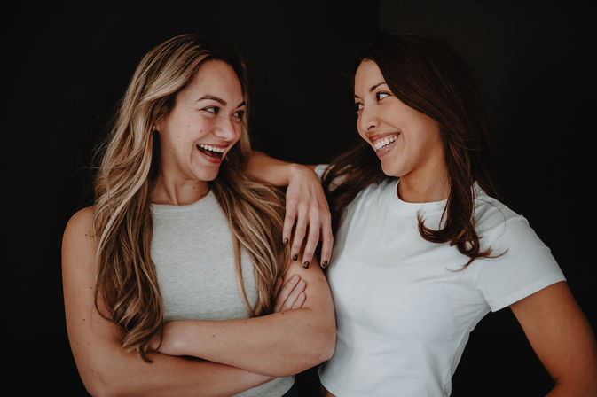 Two smiling friends — a blonde and a brunette in casual tops — laughing together in a studio portrait with one arm draped over the other against a dark background.
