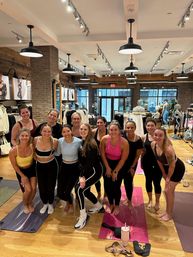 Smiling group of women in activewear posing on yoga mats after a group fitness class inside a bright urban activewear boutique with wooden floors, hanging lights and clothing racks.