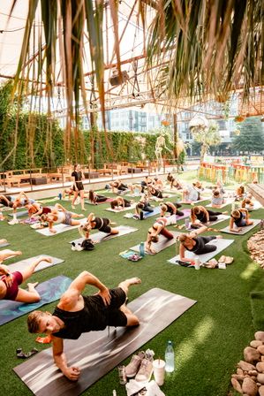 Sunlit urban outdoor yoga class on artificial turf under a shaded pergola with hanging palms and ivy, many people on mats doing side-plank stretches with city buildings in the background.