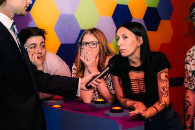 Trivia night contestants at buzzer panel — host holds a microphone to a tattooed woman answering as teammates react, set against a colorful hexagon wall.