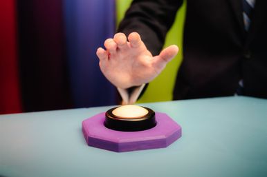 Suit-clad hand reaching to press an illuminated quiz-show buzzer on a purple octagonal base atop a bright game-show table