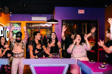 Adults and parents cheering at buzzer game stations during a neon-lit indoor arcade-style game night, colorful purple and orange walls and hanging lights creating a lively, family-friendly atmosphere.