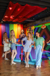 Smiling group at an indoor interactive game-show studio with rainbow curtains, colorful LED light wall, purple game podiums and a host holding a microphone.