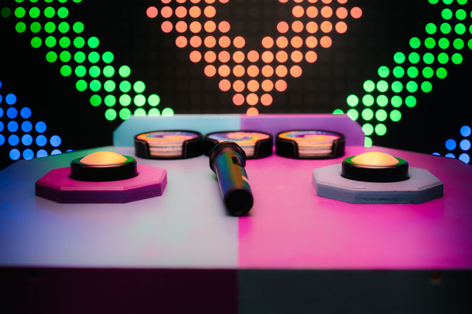 Neon-lit arcade game console with four glowing buzzer buttons and a central microphone on a pink-and-blue table, colorful LED dot wall forming arrow patterns in the background.