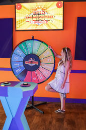 Person in a white dress spins a colorful LED-lit prize wheel at an indoor game-show attraction with bright orange walls and purple accents.