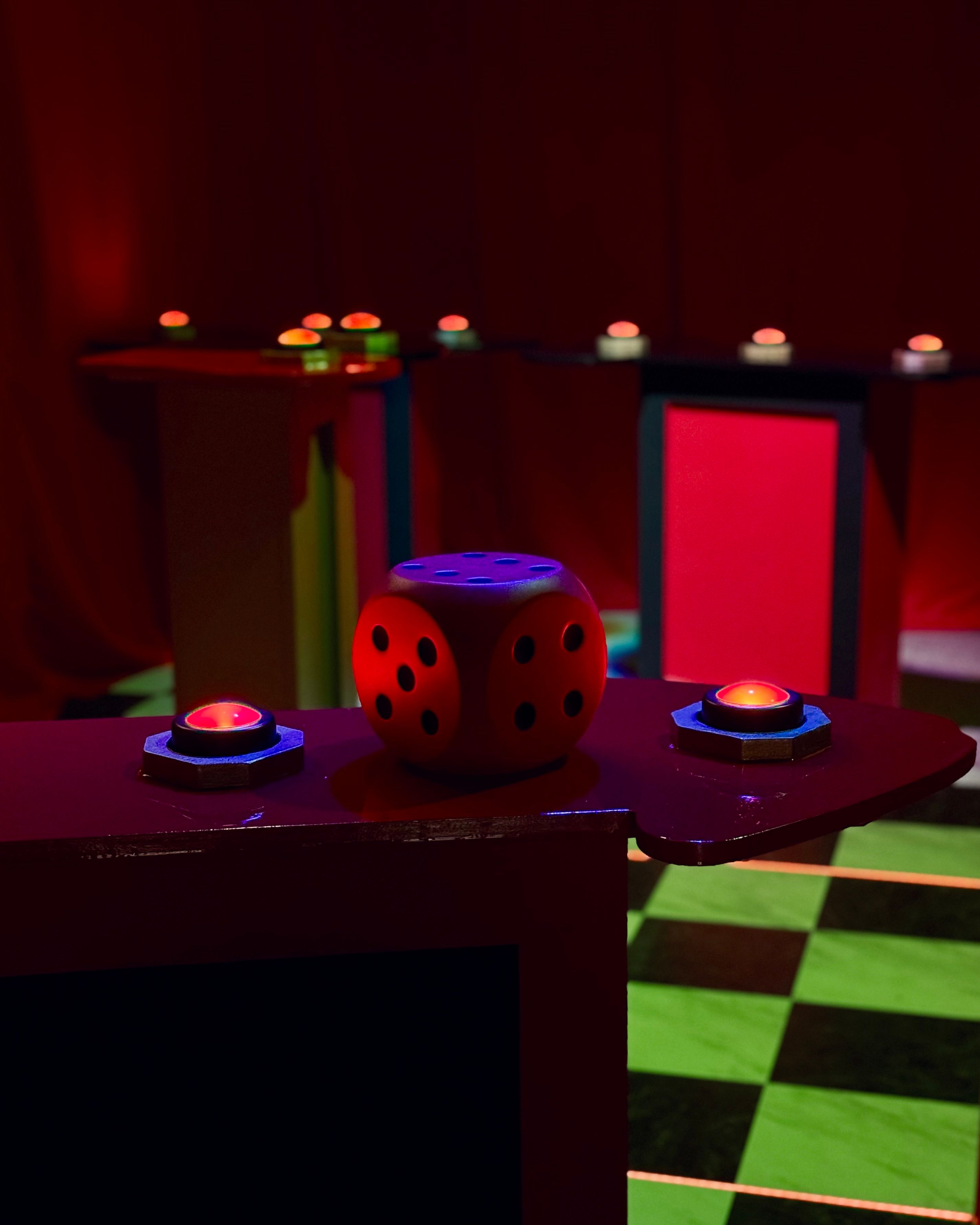 Neon-lit game show set with an oversized red die on a glossy podium, glowing red buzzer buttons and a green-and-black checkered floor.