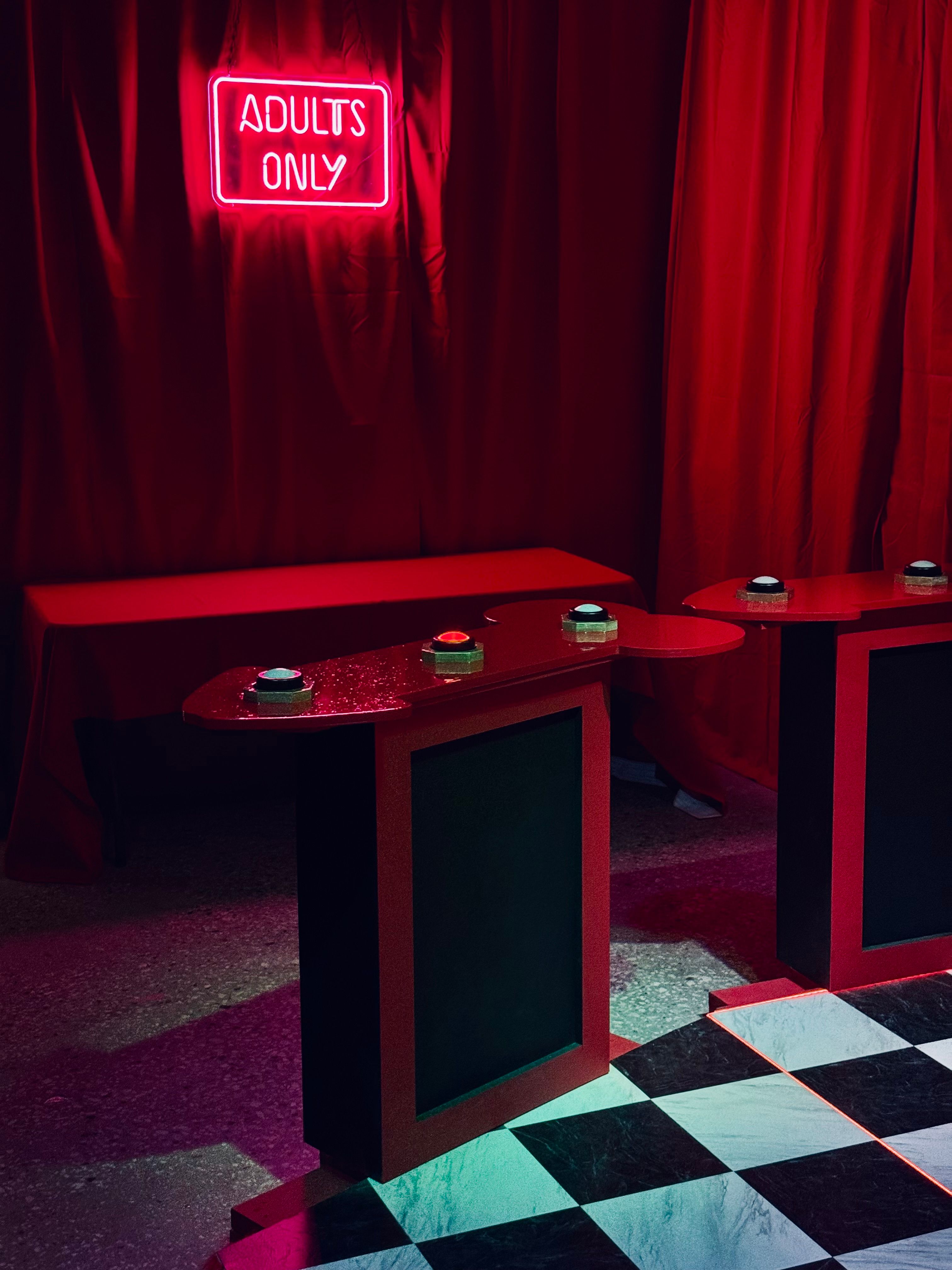 Red-lit indoor game booth with 'ADULTS ONLY' neon sign, red velvet curtains, buzzer podiums and black-and-white checkerboard floor — playful nightlife scene.
